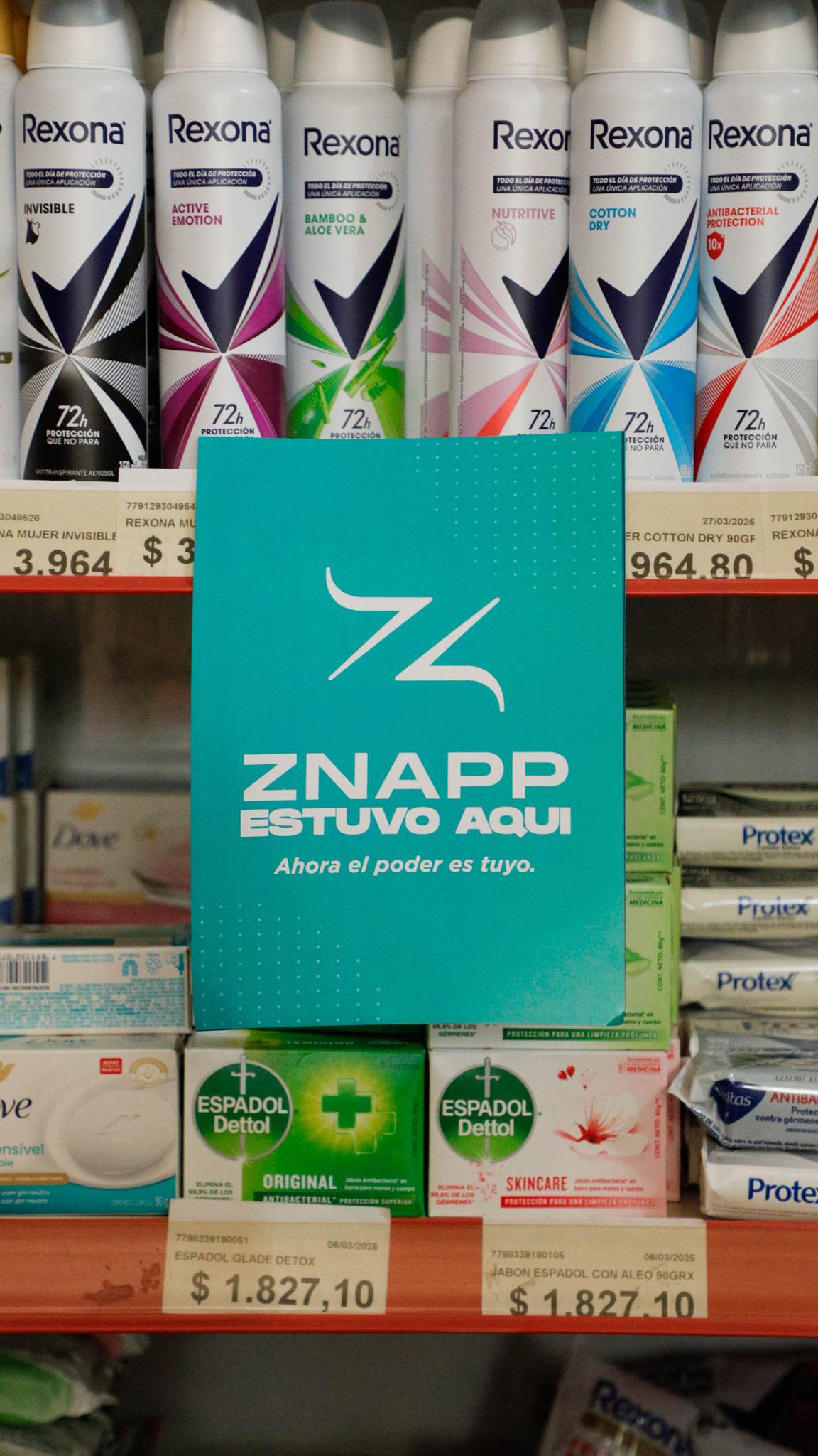 Góndola de cuidado personal con desodorantes y jabones, sus etiquetas de precio a la vista, y una tarjeta de Znapp apoyada en el estante.